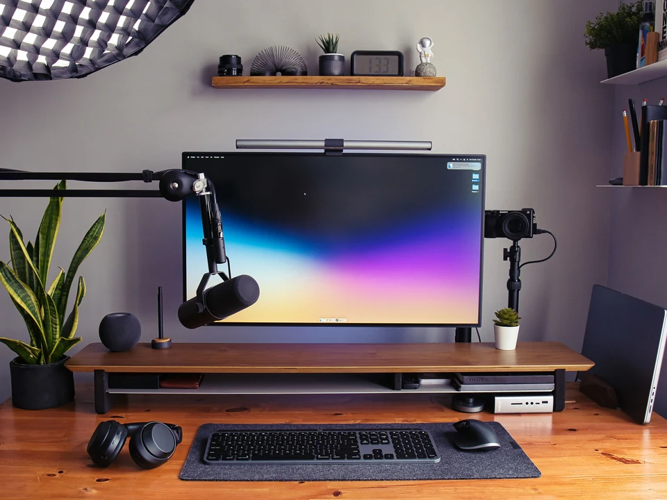 Wide desktop monitor displaying a colorful gradient above a wood desk with keyboard, microphone, and plants