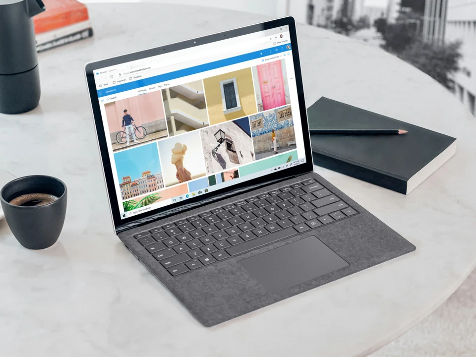 Open Microsoft Surface Laptop showing a photo gallery on a pale round table with coffee and notebook