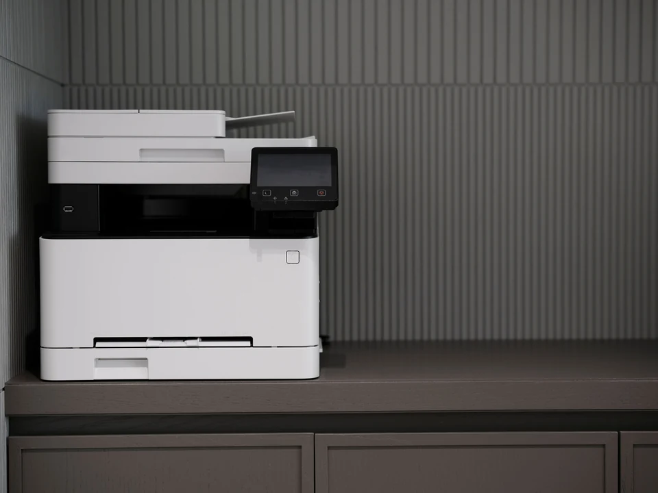 White multifunction printer on a dark cabinet against a ribbed gray office wall